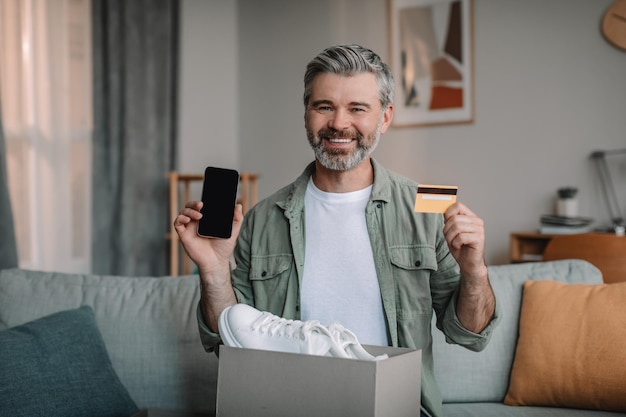 Happy elderly european male with beard unpacks cardboard box with shoes shows phone with empty