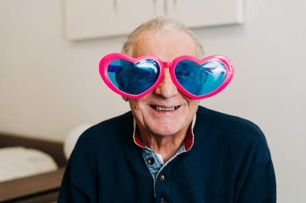 Happy elderly man portrait with heart-shaped glasses at home