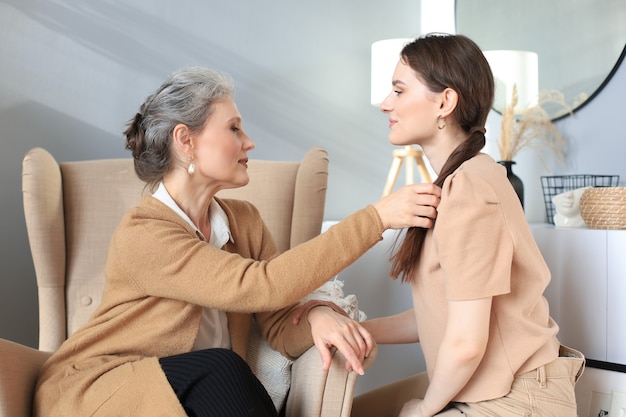 Happy elderly middle mother sitting on chair touching a strand of daughter's hair, looking each other, trusted relations. Family concept.