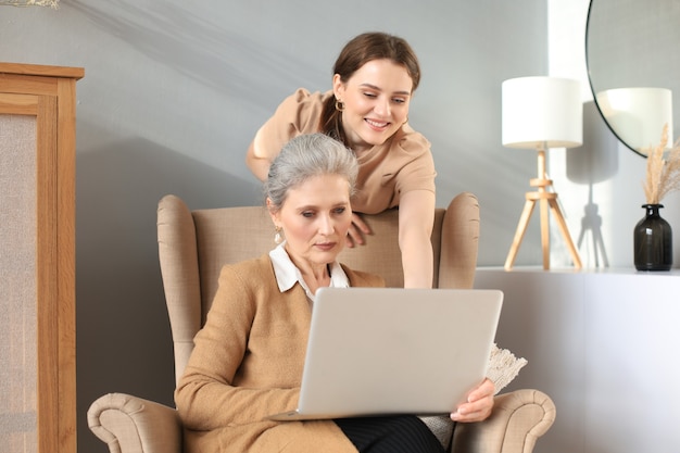 Happy elderly middle mother sitting on chair with her daughter, looking at laptop. Young woman showing video, photos to mommy, trusted relations. Family concept.