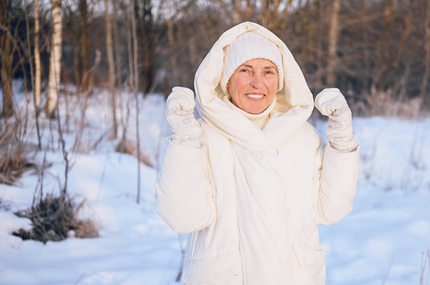 Happy elderly senior mature woman in white warm outwear playing with snow in sunny winter outdoors