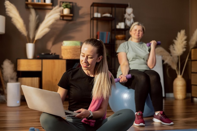 Happy elderly woman on fitball holding dumbbells in hands waiting for young helper