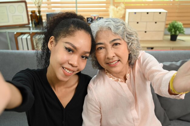 Happy elderly woman and grown up daughter having conversation by video call taking selfie while sitting in cozy living room