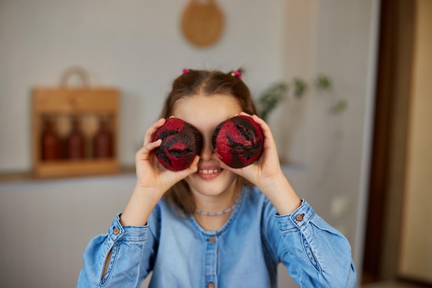 Happy emotional little girl holding cupcakes in hands covering eyes at home