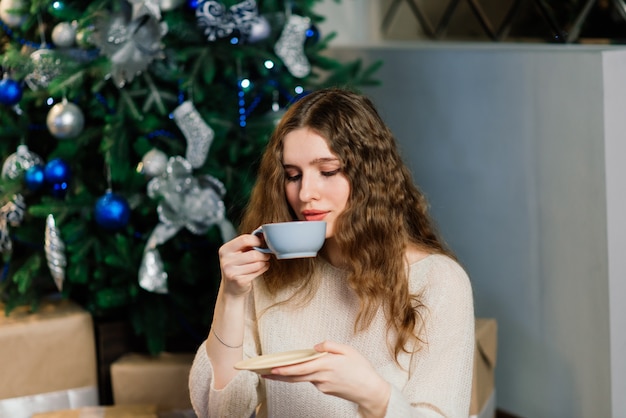 Happy emotional surprised young woman by a Christmas tree in cozy living room, concept of happiness