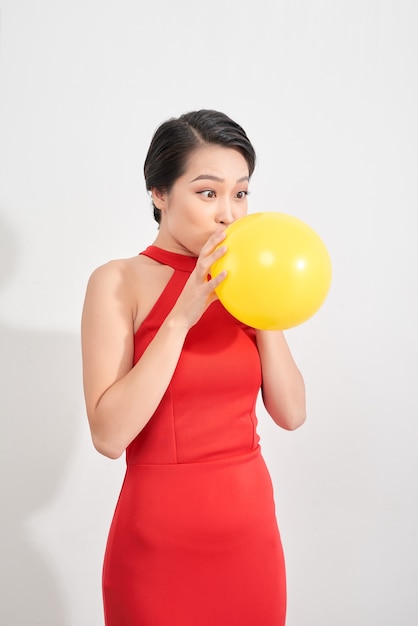 Happy emotional young asian woman in red is blowing a yellow balloon in studio. Portrait of beautiful vietnamese girl. Shopping concept