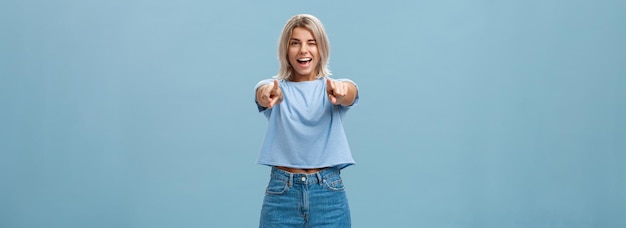 Happy enthusiastic stylish blonde female with tattoos pointing at camera as if picking or making