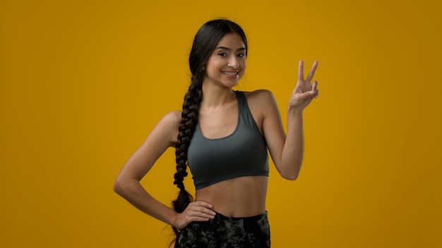 Happy ethnic Indian sport woman girl athlete in yellow studio background show victory sign peace