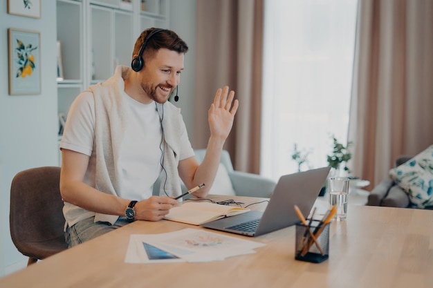 Happy European male student has online lesson waves palms and greets his tutor wears headphones modern laptop computer makes notes in notepad poses against domestic interior dressed casually