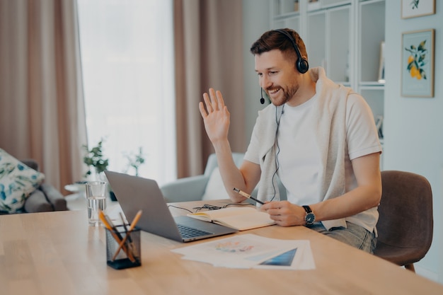 Happy European male student has online lesson waves palms and greets his tutor wears headphones modern laptop computer makes notes in notepad poses against domestic interior dressed casually