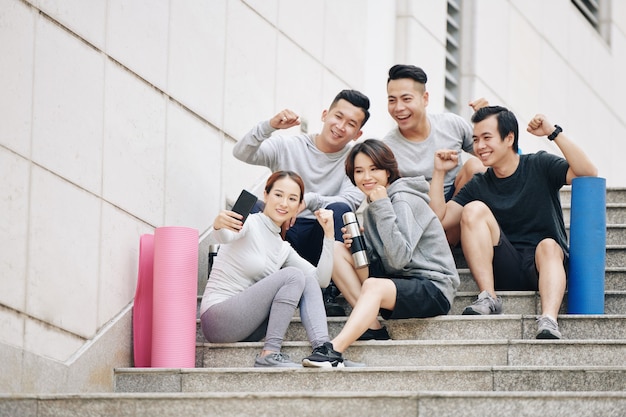 Happy excited fit friends resting on stairs and doing fist pumps when taking selfie together