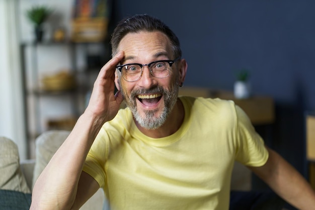 Happy excited middle aged grey haired man sitting on the sofa touching his face with a hand wearing glasses looking at camera wearing casual Thoughtful freelancer mature man working from home