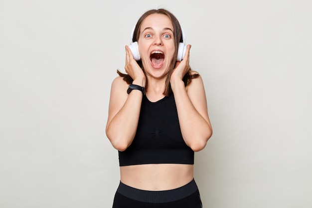 Happy excited slim woman with headphones wearing sportswear posing against gray background listening music in headphones screaming with excitement