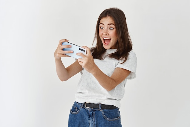 Happy and excited woman playing video game on smartphone, standing in t-shirt against white wall.