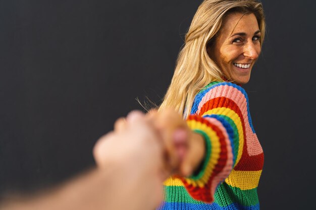 Photo happy excited woman pulling hand wearing colorful rainbow sweater standing over black background lgbt celebrating concept image