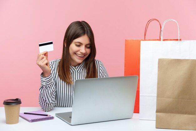 Happy excited woman sitting at laptop and holding credit card, doing shopping online, paper bags lying on desk, fast delivery service. Indoor studio shot isolated on pink background