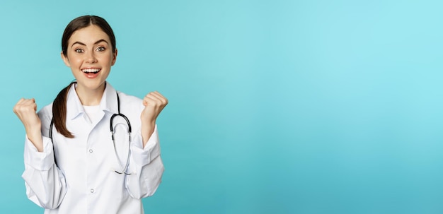 Photo happy and excited young female doctor physician celebrating achieve goal and smiling pleased winning standing in white coat against blue background