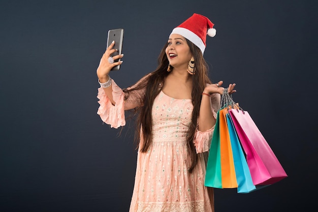 Happy excited young girl in a Santa Claus hat holding shopping bags and showing screen of smartphone or using mobile phone or tablet over gray background Christmas concept