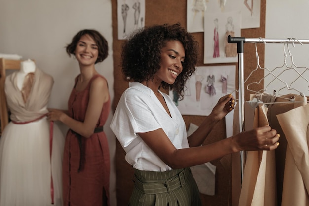 Happy excited young women in trendy summer outfits smile work as fashion designers in cozy office