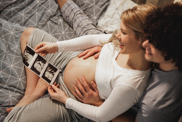 Photo happy expecting couple looking at medical scan of their baby while relaxing on a bed in bedroom.