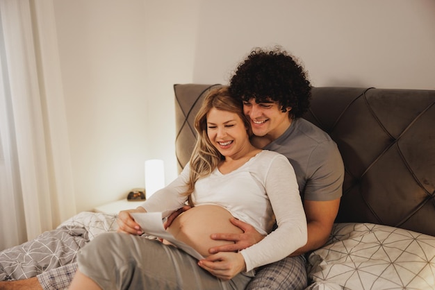 Happy expecting couple looking at medical scan of their baby while relaxing on a bed in bedroom.