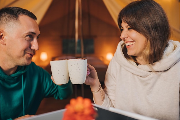 Happy family couple freelancers drink coffee while working laptop on cozy glamping tent in summer evening Luxury camping tent for outdoor holiday and vacation Lifestyle concept