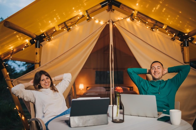 Happy family couple freelancers working laptop on a cozy glamping tent in summer evening Luxury camping tent for outdoor holiday and vacation Lifestyle concept