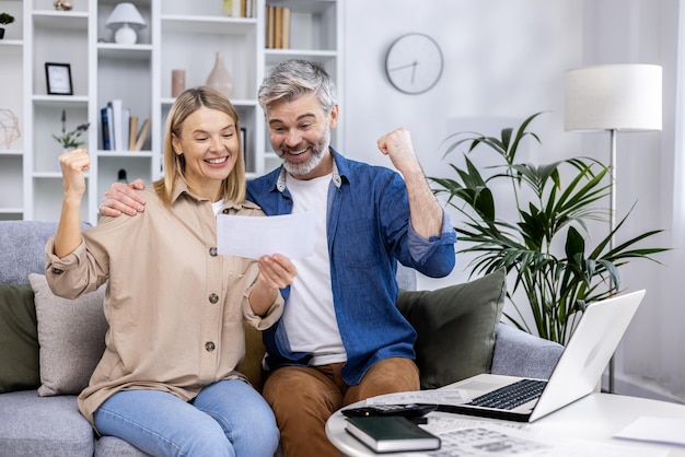 Happy family couple mature adult man and woman sitting at home on sofa celebrating victory holding