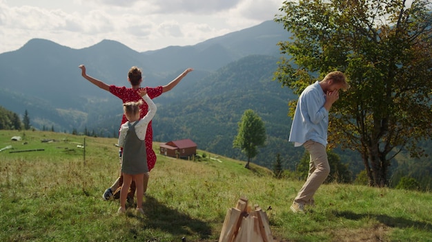 Happy family funny dancing on green mountain hill Joyful young parents playing active game with children on summer nature Cheerful people have fun spending vacation with kids on sunny valley