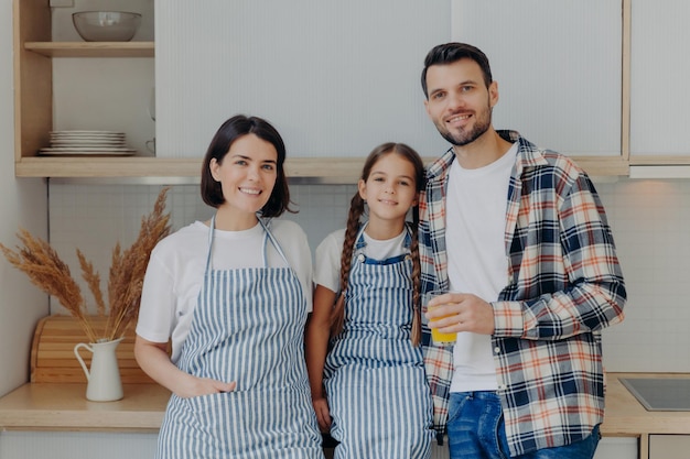 Happy family have great time together pose in modern kitchen at home Glad man in checkered shirt holds glass of juice small child with pigtails pretty housewife in apron Girl with parents