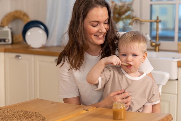 Happy family at home mother feeding baby in kitchen little boy with messy funny face eats healthy fo