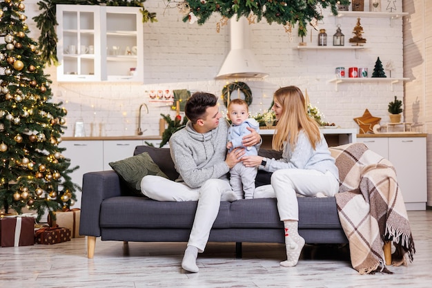 A happy family is sitting on the couch with their little son near the Christmas tree and smiling joyfully Home festive atmosphere of the new year