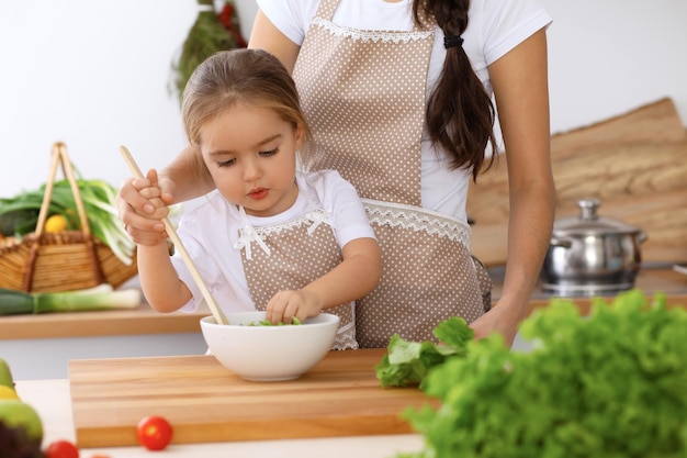 Happy family in the kitchen Mother and child daughter cooking tasty breakfest of fresh salad Little helper slicing and mix tomatos and greenery