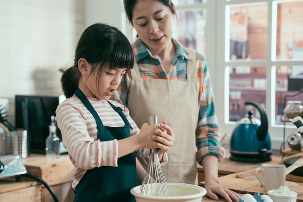 happy family in kitchen. young asian mother and child daughter preparing dough bake cookies for birthday. adorable kid with mom mix flour and egg together in bowl at home. elegant woman look at girl