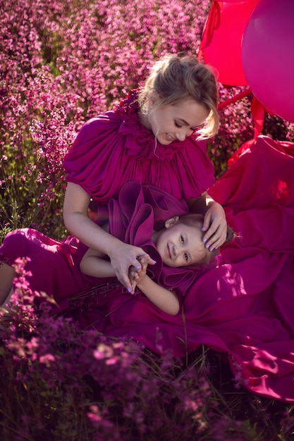 Happy family mom and daughter in pink dresses are sit in a field with flowers and big balloons at sunset in summer