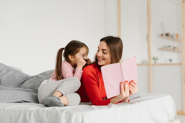 Happy family mother and child daughter reading holding book lying in bed