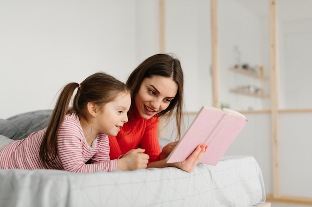 Happy family mother and child daughter reading holding book lying in bed