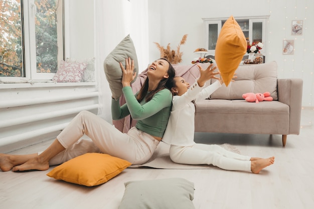 A happy family! Mother and her little daughter girl fight pillows on the floor near the sofa. Happy family games.