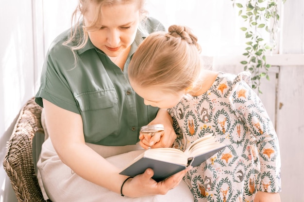 Happy family mother with cute little kid daughter girl have fun together in suburb countryside home on sunny summer day candid mom hugging and reads book to child exploring book with magnify glass