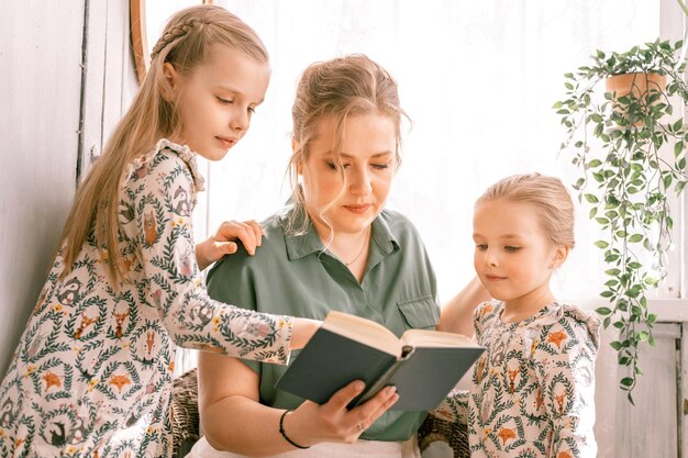 Happy family mother with cute little kids daughters girls have fun together in suburb countryside home on sunny summer day candid mom hugging and reads book to children people love and care