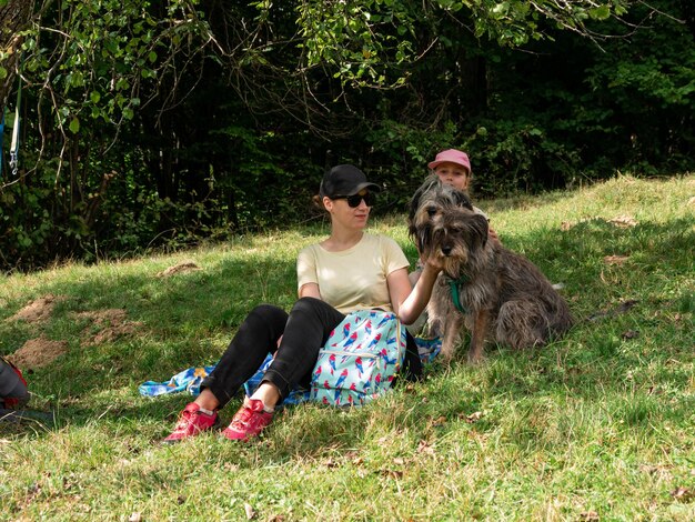 Happy family mother with daughter and two dogs sitting on green grass lawn having fun picnic spending time together hiking in the mountains People and pets outdoors lifestyle photo candid emotions