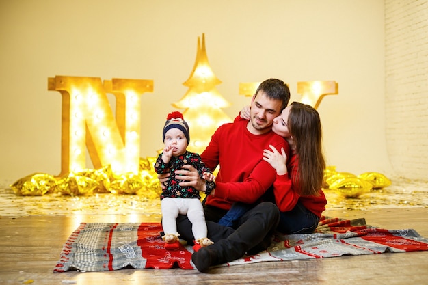 A happy family in red sweaters is sitting together on the floor. Christmas holiday atmosphere. A child in a santa costume. Family relationship concept