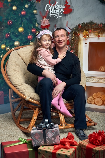 Happy family spends time together on winter vacation at home by the fireplace near the Christmas tree with gifts. Cute little girl with her father in the chair at the Christmas tree.