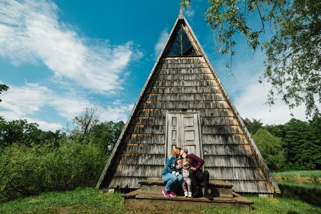 Happy family on vacation by the lake in nature Young mother and father with two children on the background of an old wooden house Walks in the park