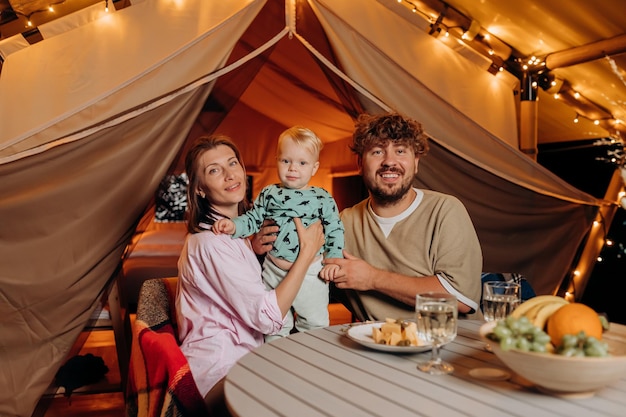 Happy family with lovely baby have dinner and spend time together in glamping on summer evening near cozy bonfire Luxury camping tent for outdoor recreation and recreation Lifestyle concept