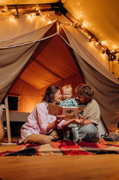 Happy family with lovely baby playing and read book together in glamping on summer evening Luxury camping tent for outdoor recreation and recreation Lifestyle concept