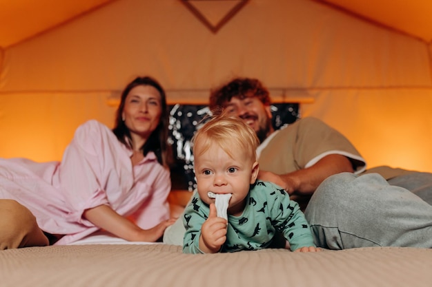 Happy family with lovely baby playing and spend time together in cozy glamping on summer evening while lying on bed Luxury camping tent for outdoor recreation and recreation