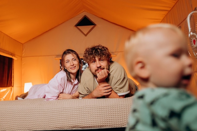 Happy family with lovely baby playing and spend time together in cozy glamping on summer evening while lying on bed Luxury camping tent for outdoor recreation and recreation