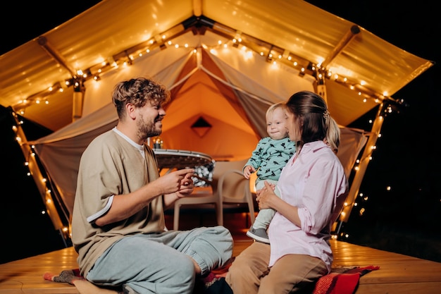 Happy family with lovely baby relaxing and spend time together in glamping on summer evening near cozy bonfire Luxury camping tent for outdoor recreation and recreation Lifestyle concept