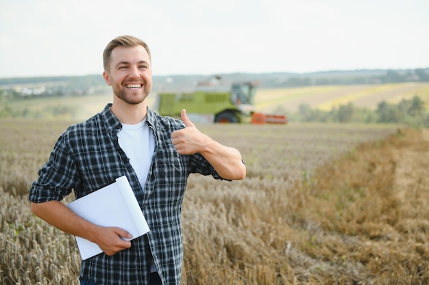 Happy farmer proudly standing in a field Combine harvester driver going to crop rich wheat harvest Agronomist wearing flannel shirt looking at camera on a farmland